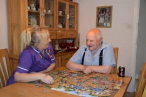 A caregiver from Pride Homecare engaging with an elderly client in a warm, home environment, sharing a smile over a jigsaw puzzle, exemplifying the compassionate care provided.
